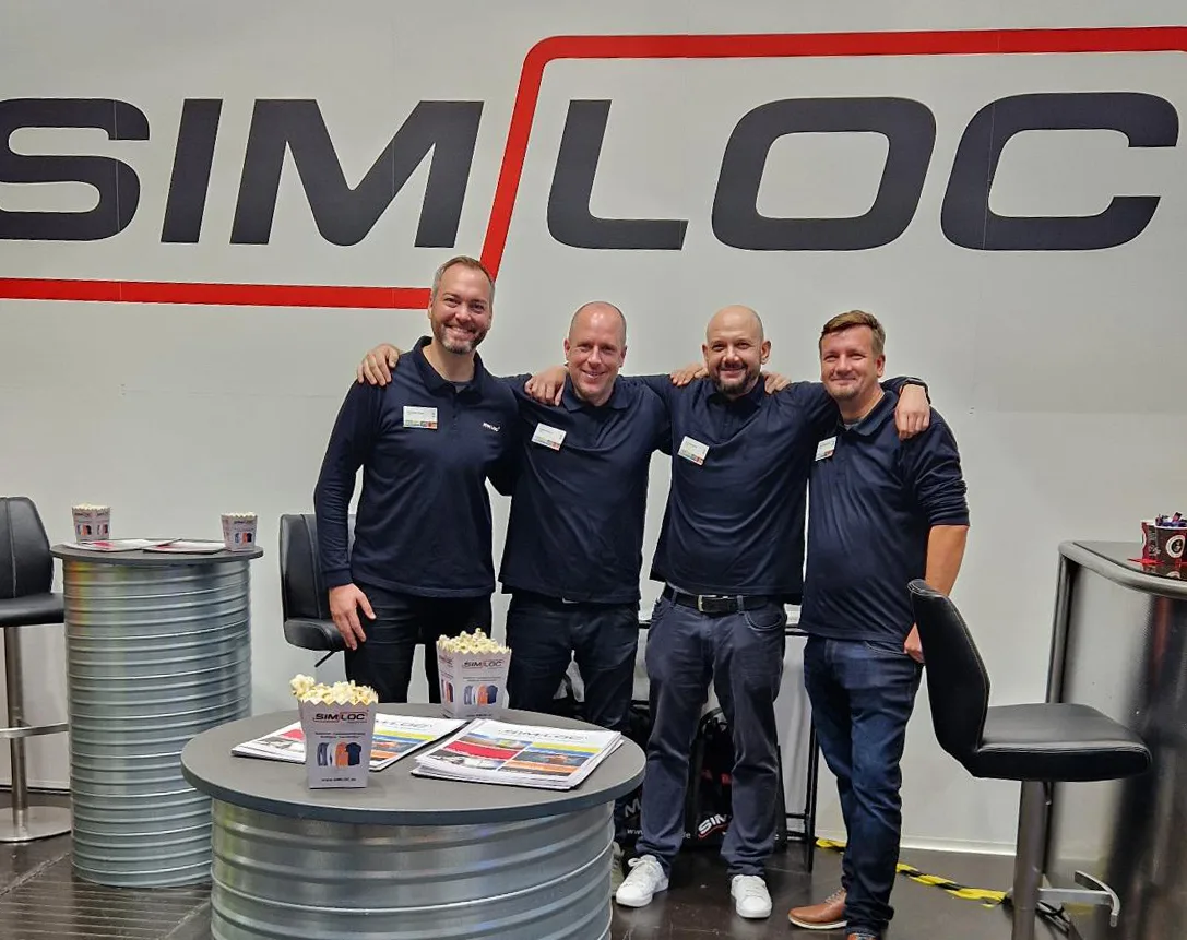 Vier Männer im blauen Shirt präsentieren lachend SIMLOC-Produkte und Broschüren, die innovative Sicherheitslösungen zeigen.